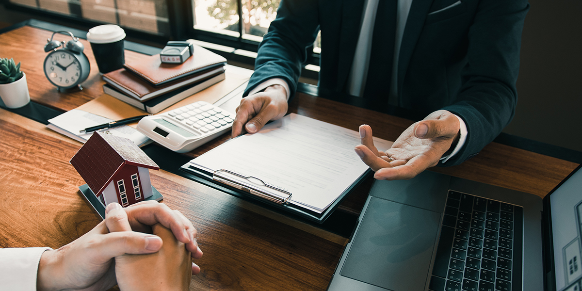 Mortgage broker discussing home loan options with client, featuring a model house, paperwork, calculator, and laptop on a desk.