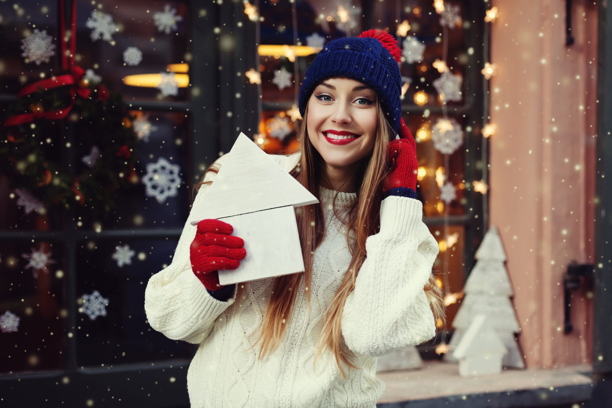 Young woman in cozy winter attire holding a decorative house-shaped gift, smiling in front of a festive holiday backdrop with snowflakes and Christmas decorations, representing the theme of mortgage refinancing as a holiday gift.