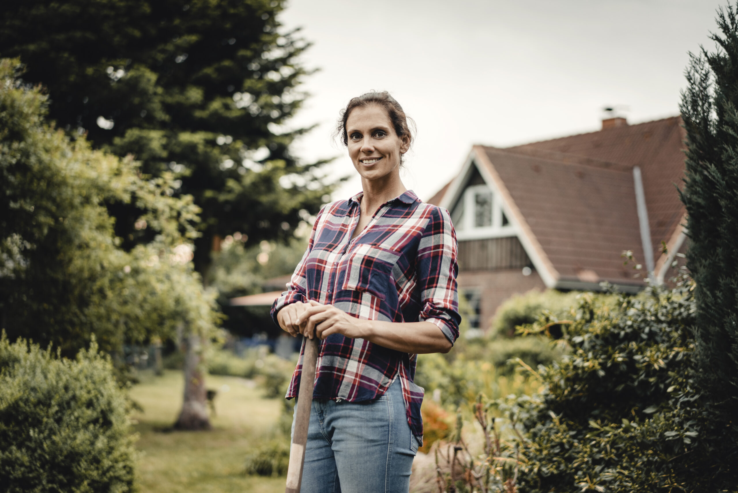 Woman in a plaid shirt standing in a garden, holding a gardening tool, with a house and greenery in the background, illustrating homeownership and outdoor living.