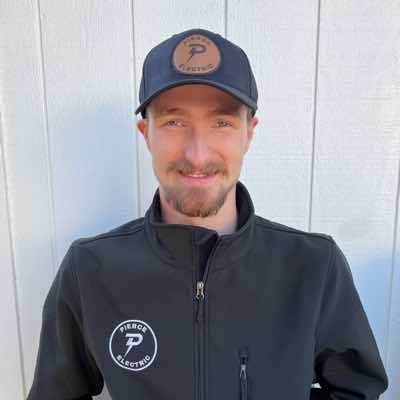Man wearing a black jacket with Pierce Electric logo and a matching cap, smiling in front of a light-colored wooden background.